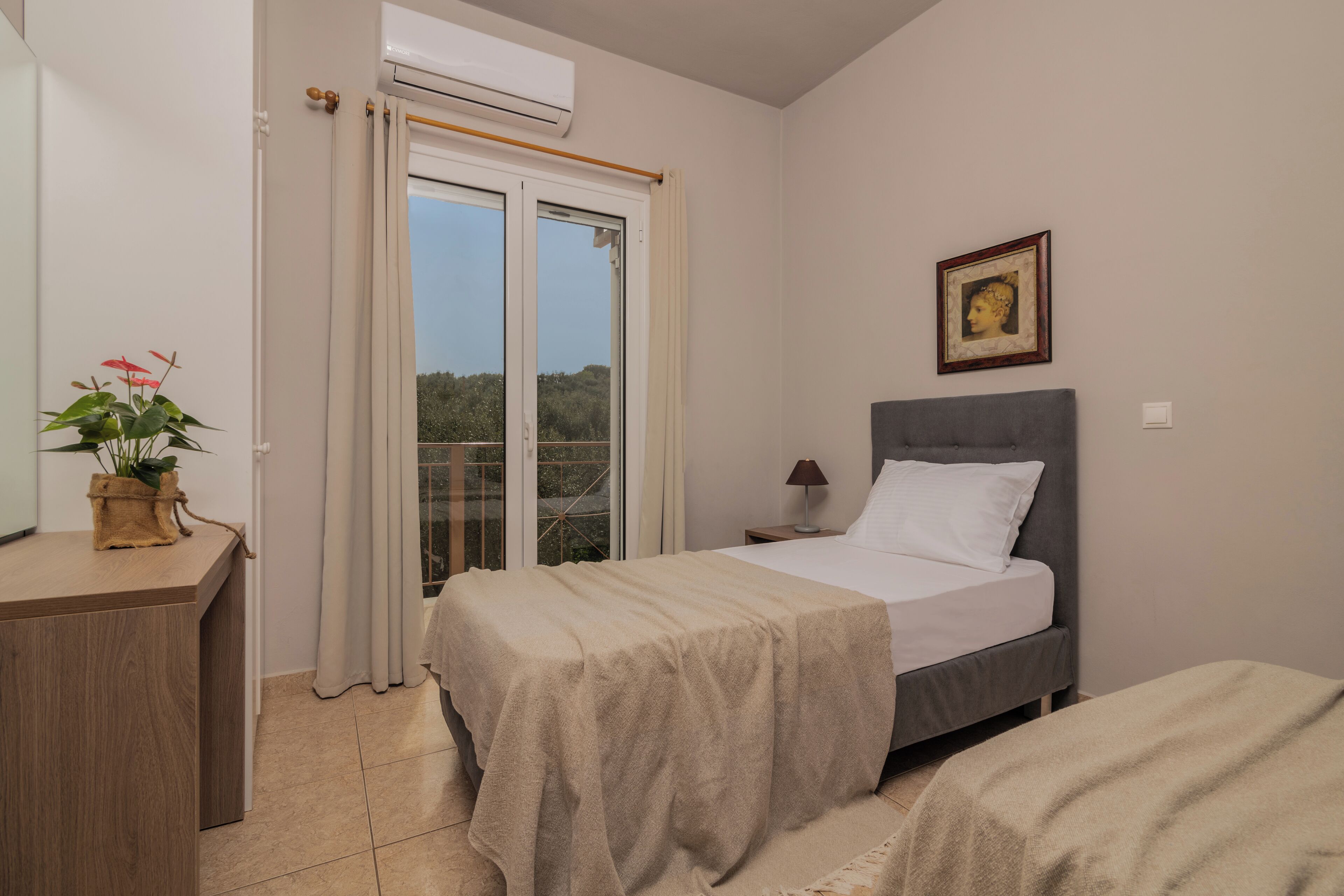 A bedroom with a bed, a bedside table, a lamp, a framed picture, a window with curtains, and a potted plant.
