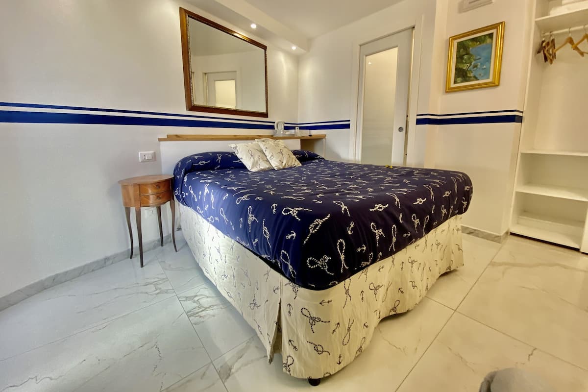 A bedroom with a bed, a nightstand, a mirror, a door, and a shelf with a picture.
