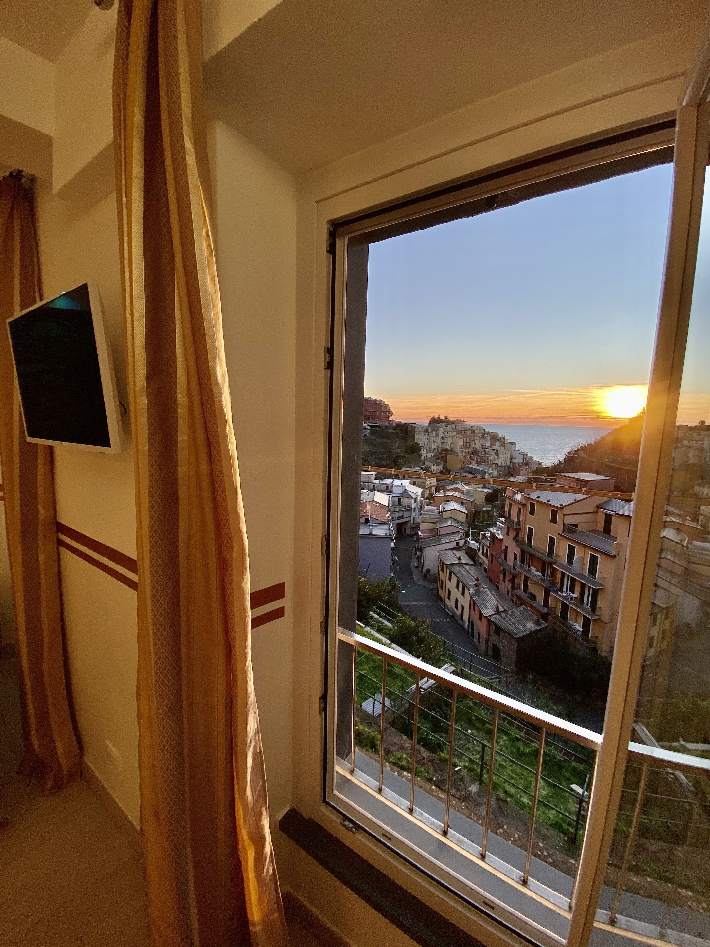 A room with a large window overlooking a coastal town at sunset.