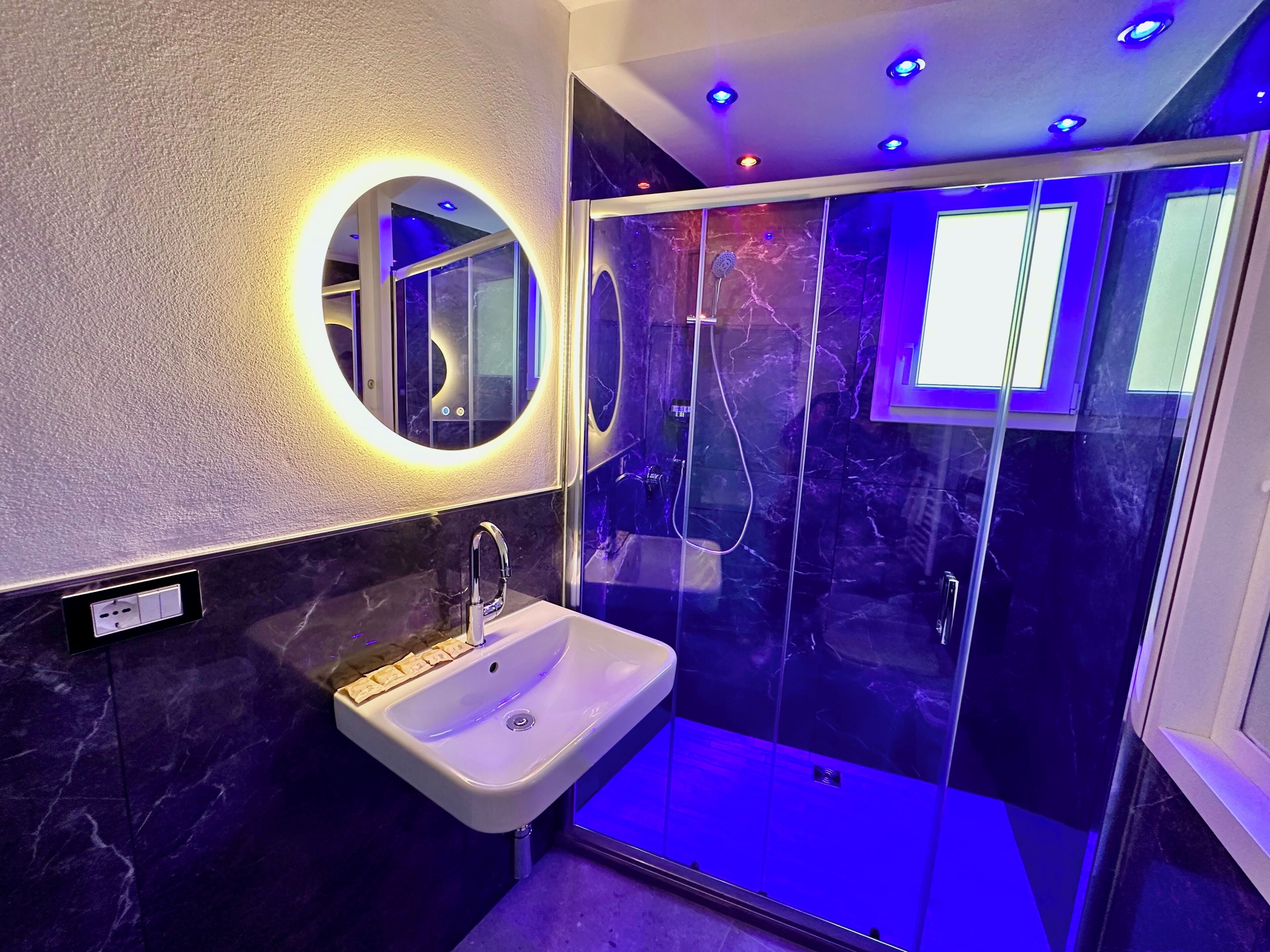 A modern bathroom with a marble sink, a glass shower enclosure, and a circular mirror with underglow lighting.