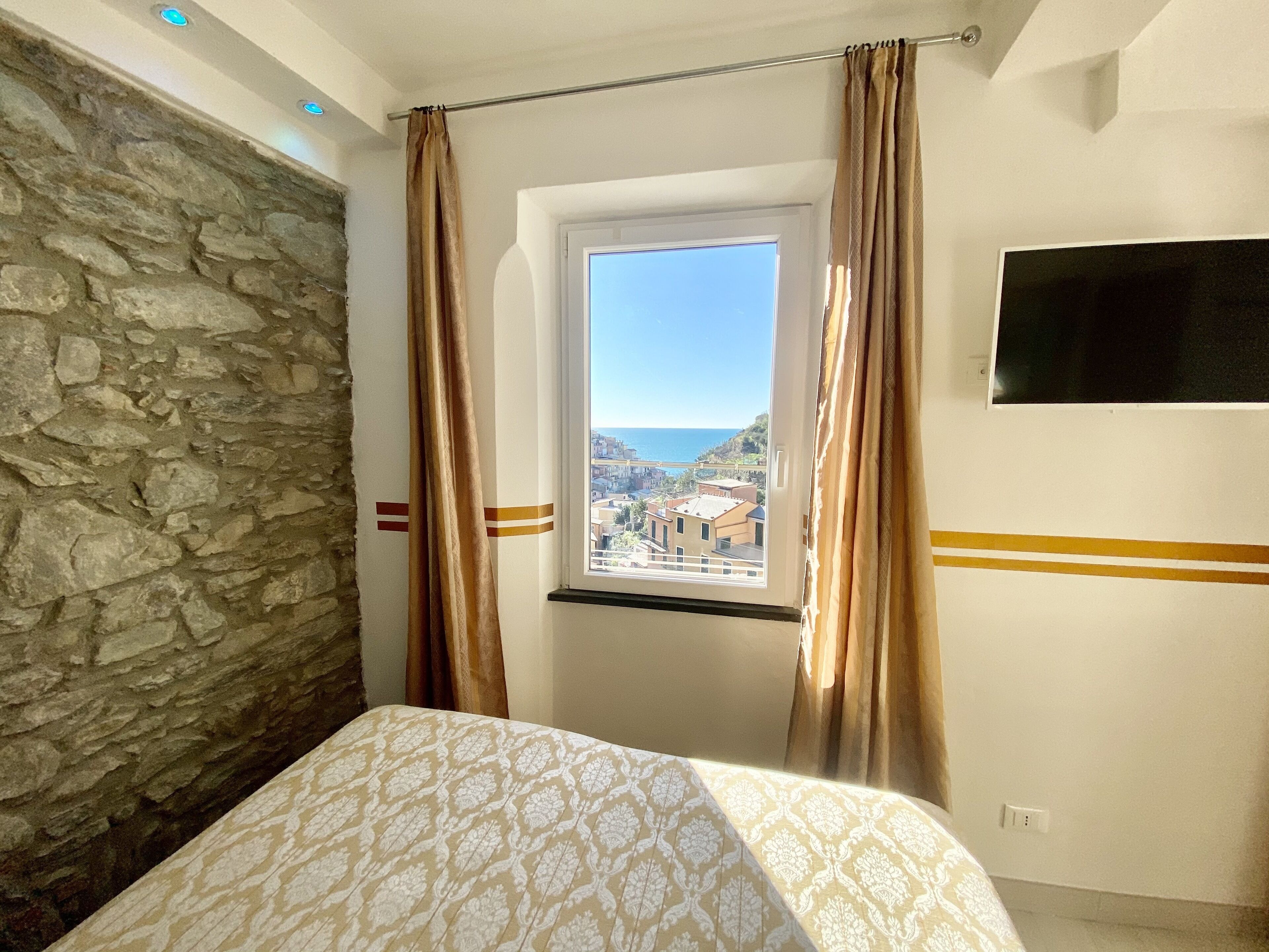 A bedroom with a stone wall, a window with curtains, and a bed with a patterned bedspread.