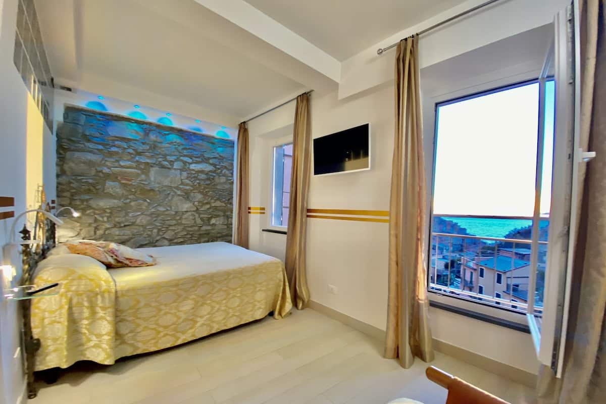 A bedroom with a stone wall, a bed with a patterned bedspread, a TV on the wall, and a window with a view of buildings and water.