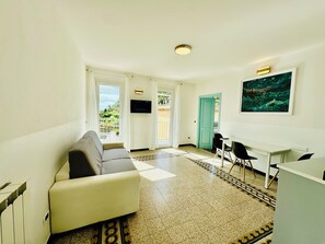 A living room with a sofa, a TV, a desk with chairs, and a door leading to an outdoor area.