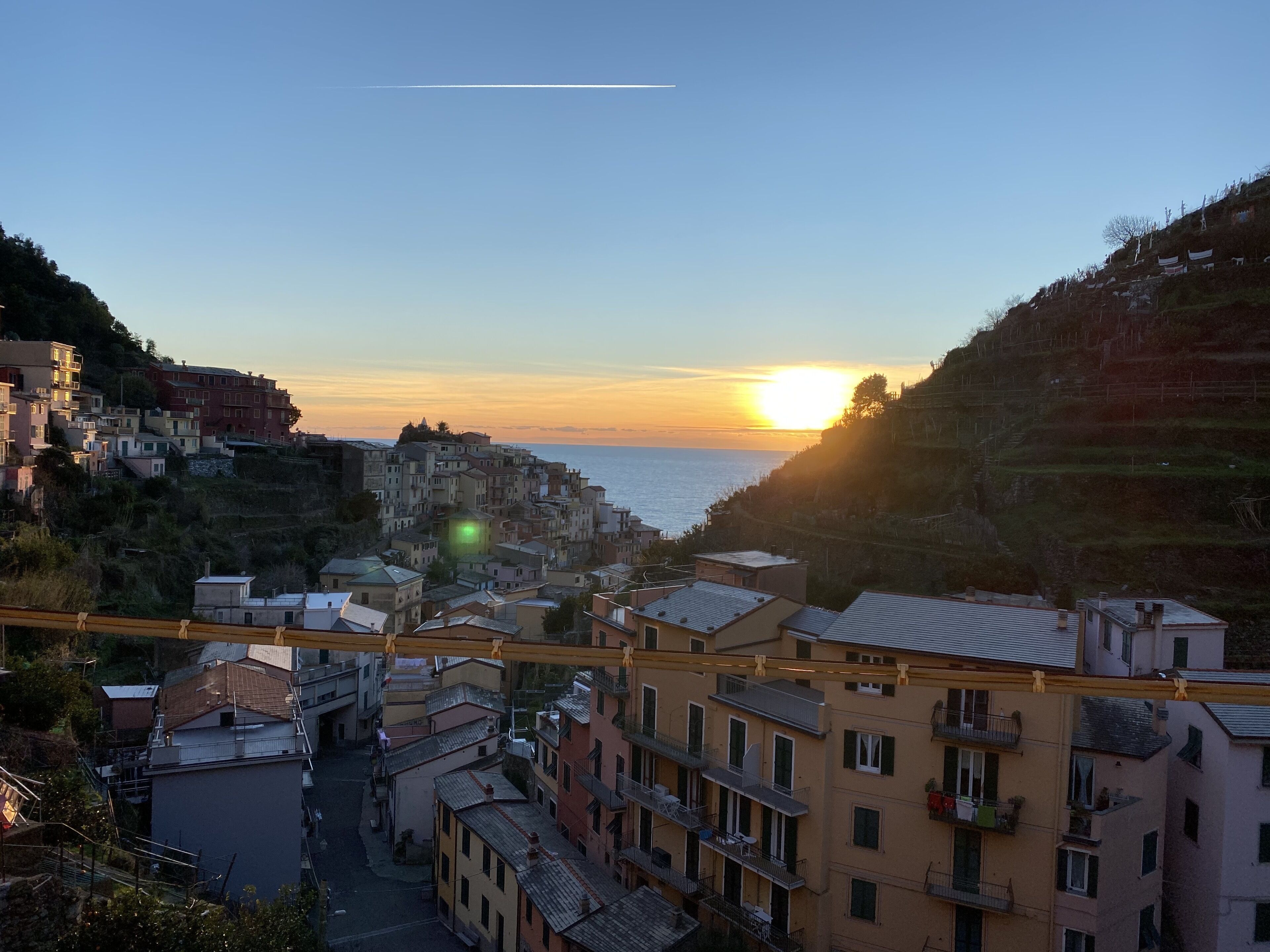 A coastal town with buildings along the hillside, a clear sky, and the sun setting over the sea.