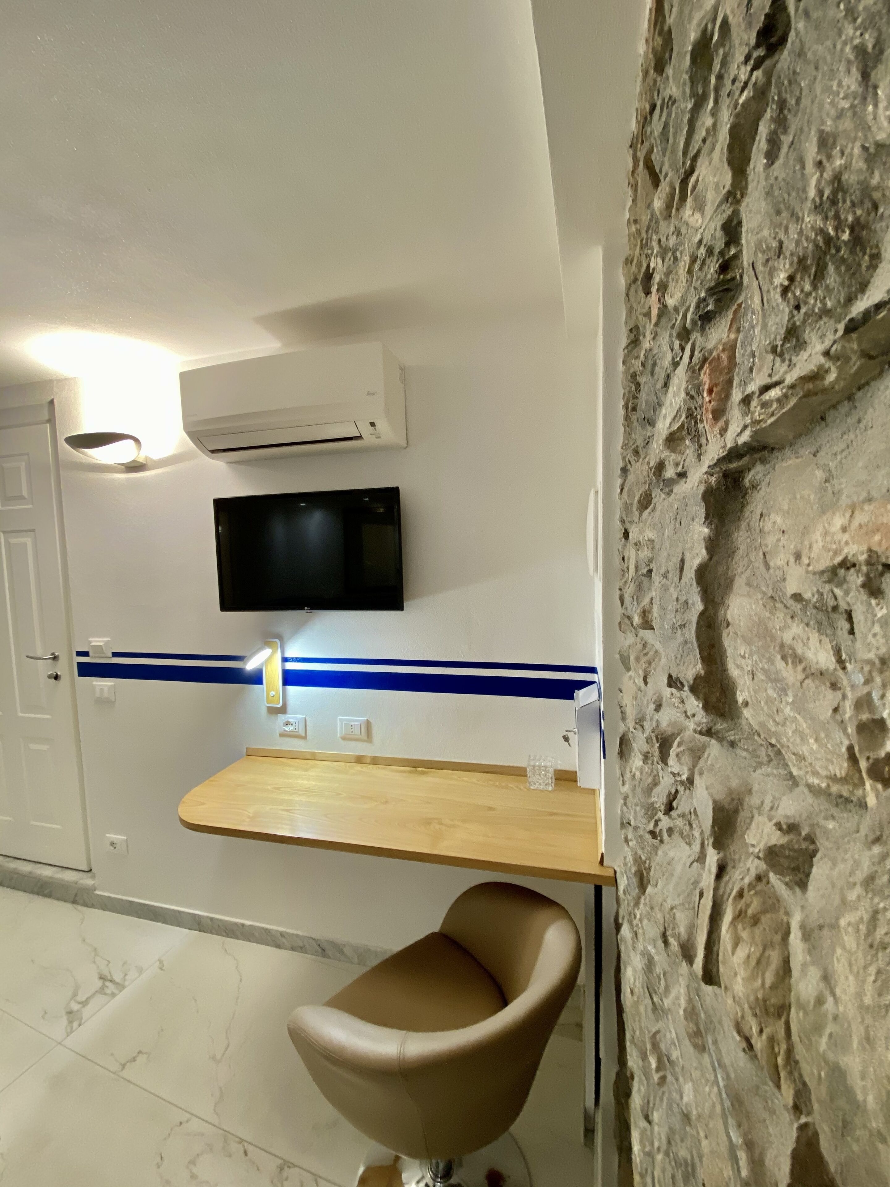 Een moderne badkamer met een accentmuur van steen, een wandairconditioner, een flatscreen-televisie, een houten wastafelmeubel en een moderne stoel.