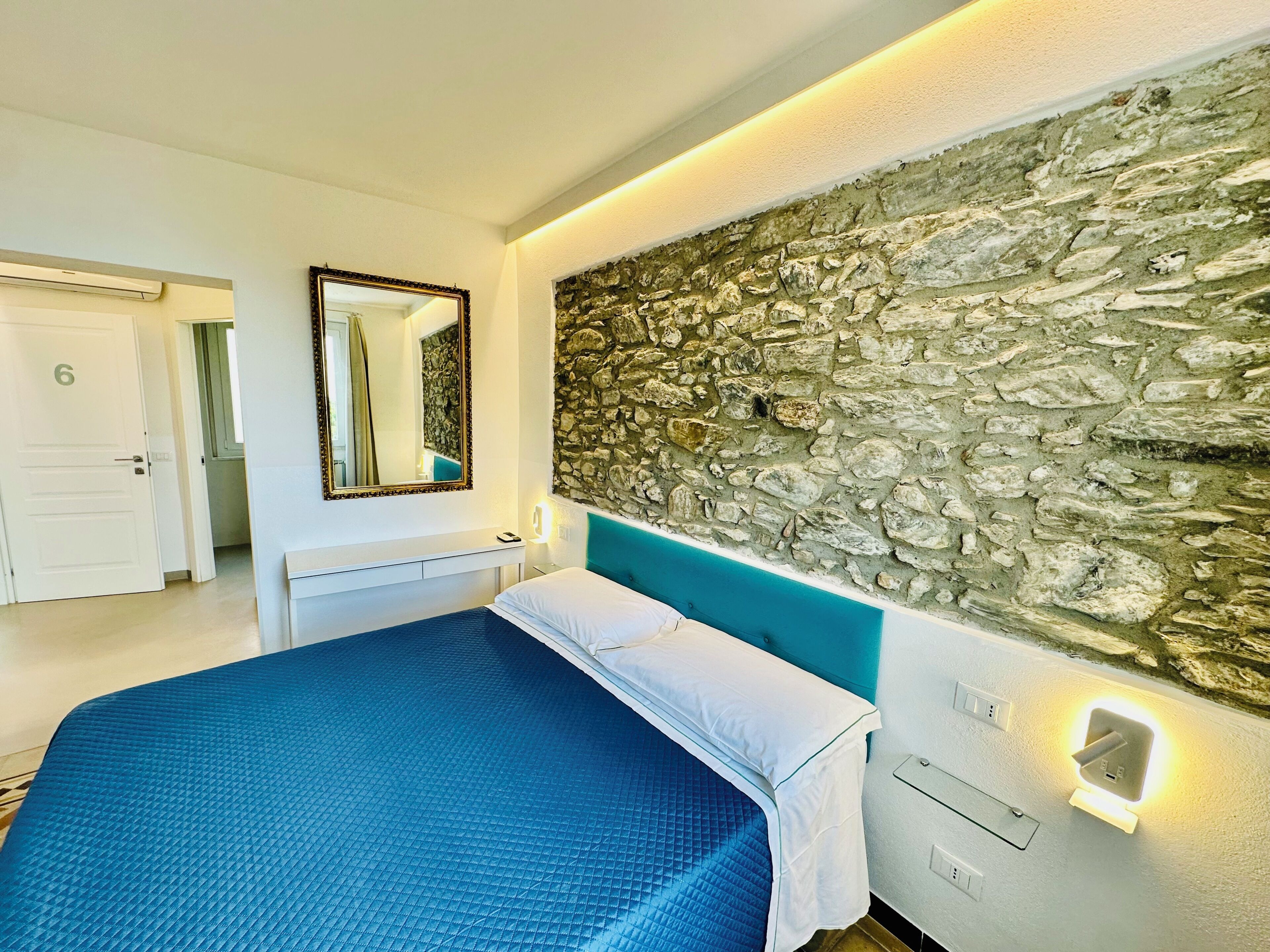 A bedroom with a stone wall, a bed with a blue bedspread, a white bedside table, a mirror, and a door labeled with the number 6.