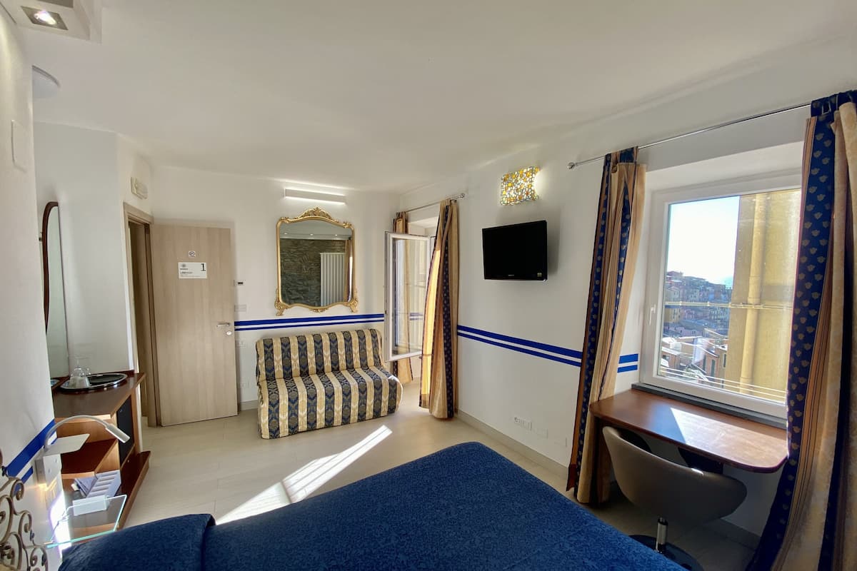 A hotel room with a bed, a desk with a chair, a TV, a sofa, and a window with curtains.