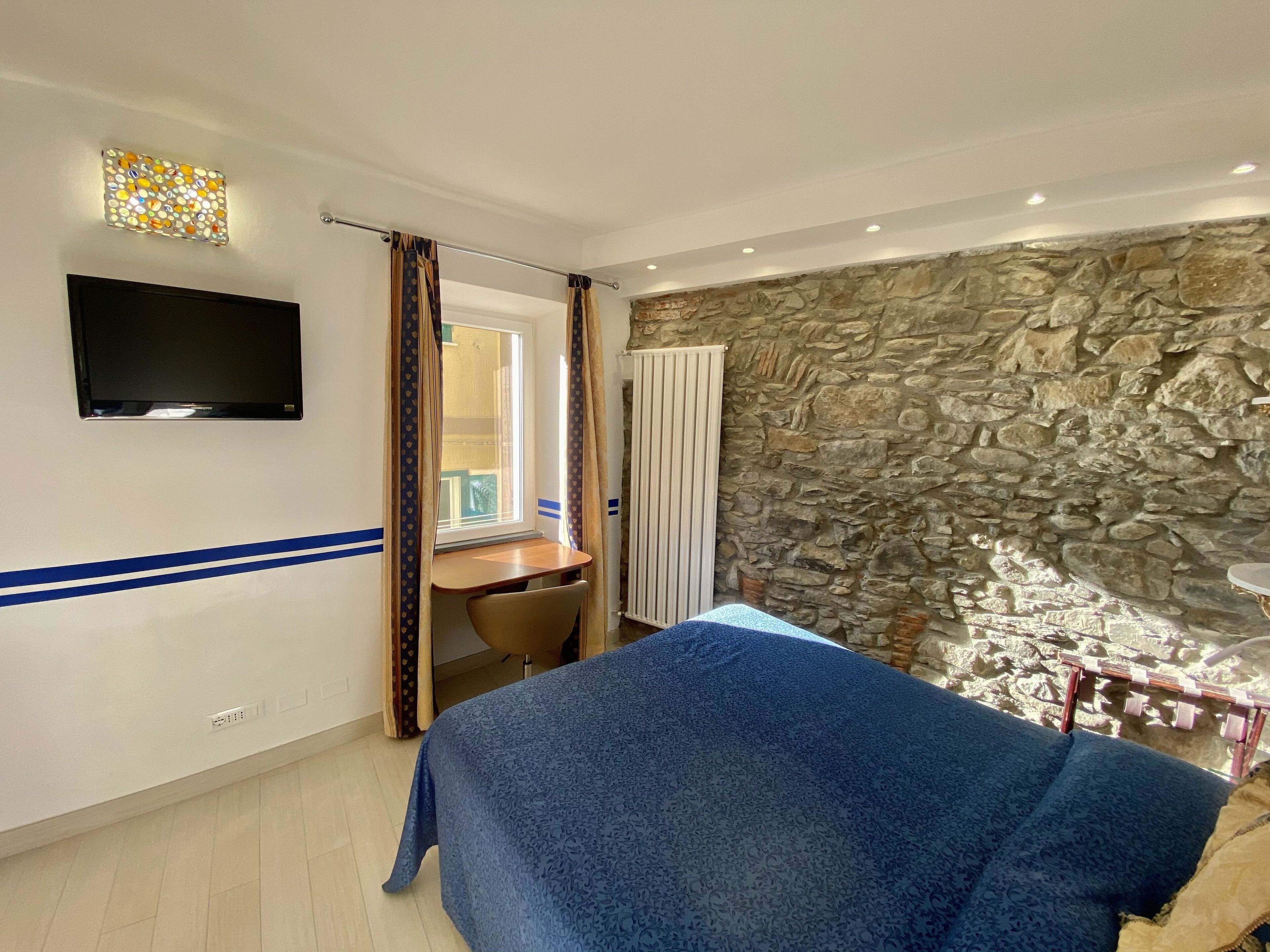A bedroom with a bed, a desk, a television, a stone wall, and a window.