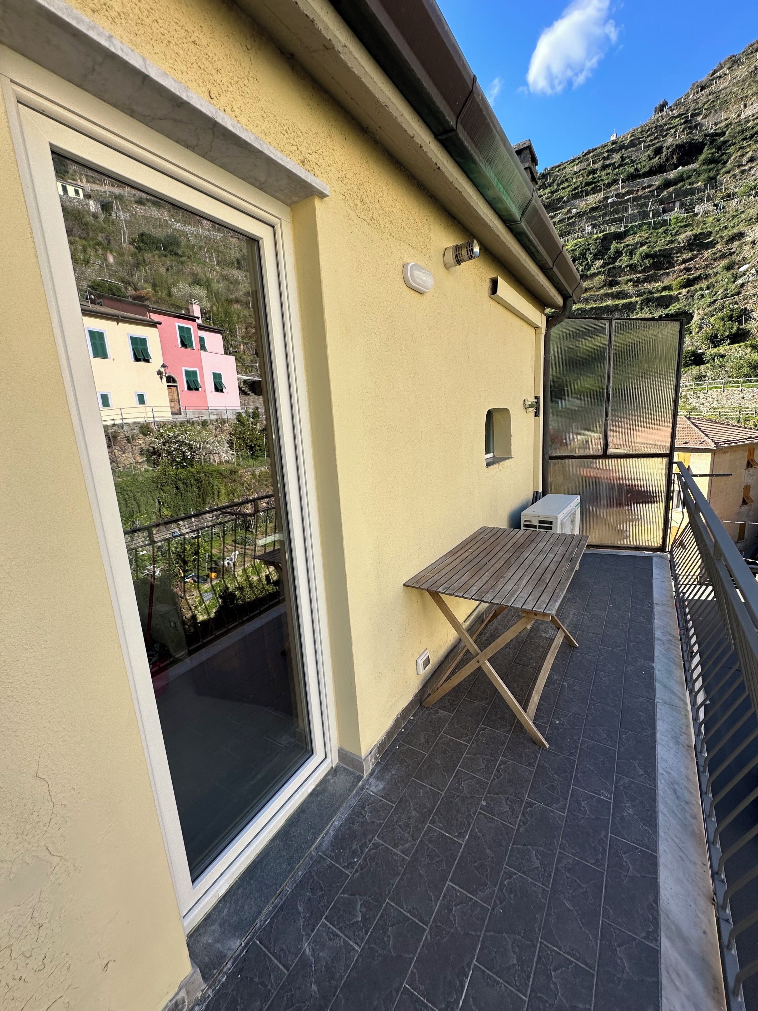 A balcony with a table and chairs, a sliding glass door, and a view of a hillside with buildings.