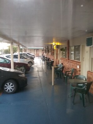 A covered outdoor area with several cars parked on the left, a row of tables and chairs on the right, and a building with a number '2' on the door.