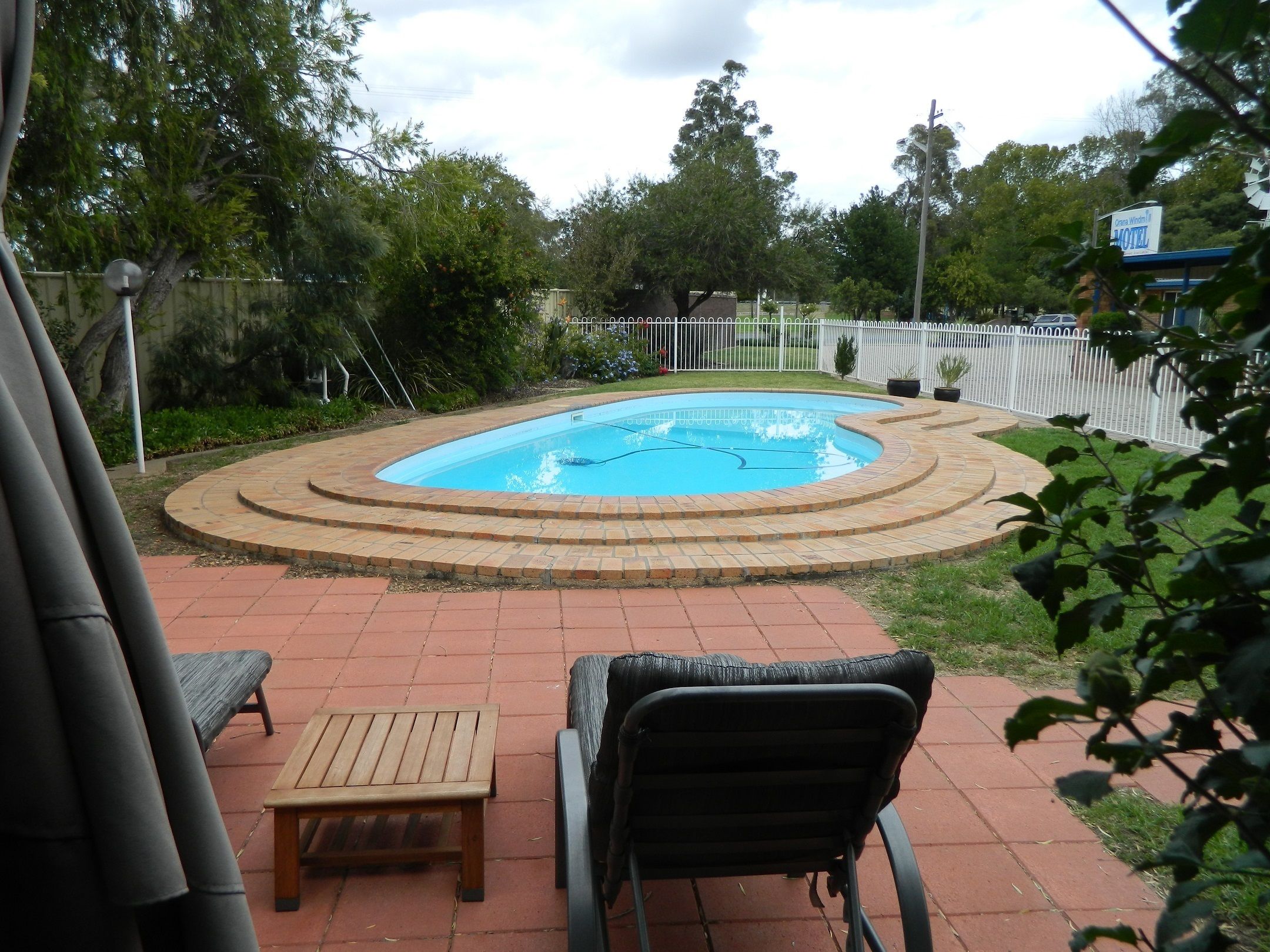 Una piscina con borde de ladrillo, rodeada por una valla blanca, y un banco y una silla al frente.