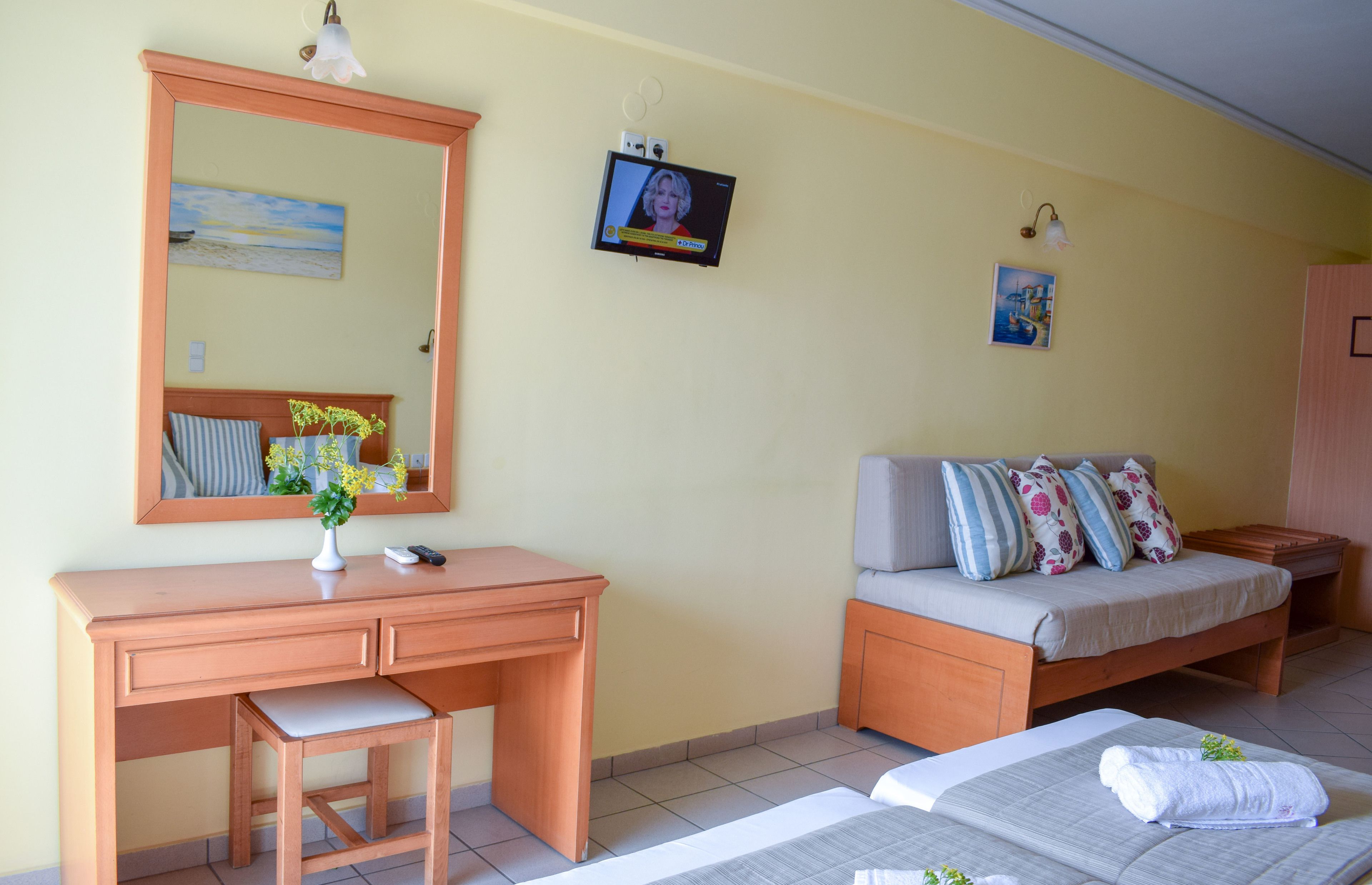Una habitación con escritorio de madera, cama con almohadillas, mesita de noche pequeña y televisor instalado en la pared.
