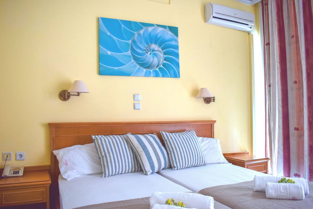 A double bed with striped pillows, a wooden headboard, two bedside lamps, a wall-mounted air conditioner, and a painting of a blue spiral.