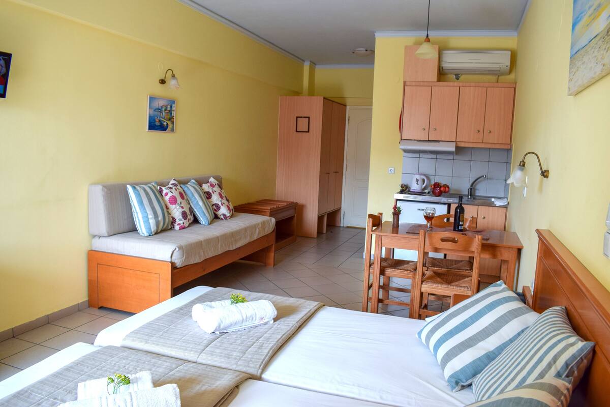A hotel room with two beds, a sofa, a dining table, and a kitchen area.