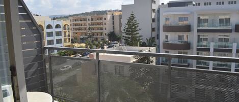 A balcony with a mesh railing, a round table, and a white chair. In the background, there are multi-story buildings with balconies and a clear sky.
