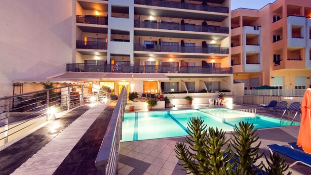 Un hotel de varios pisos con piscina, asientos exteriores e iluminación destacada en la entrada.