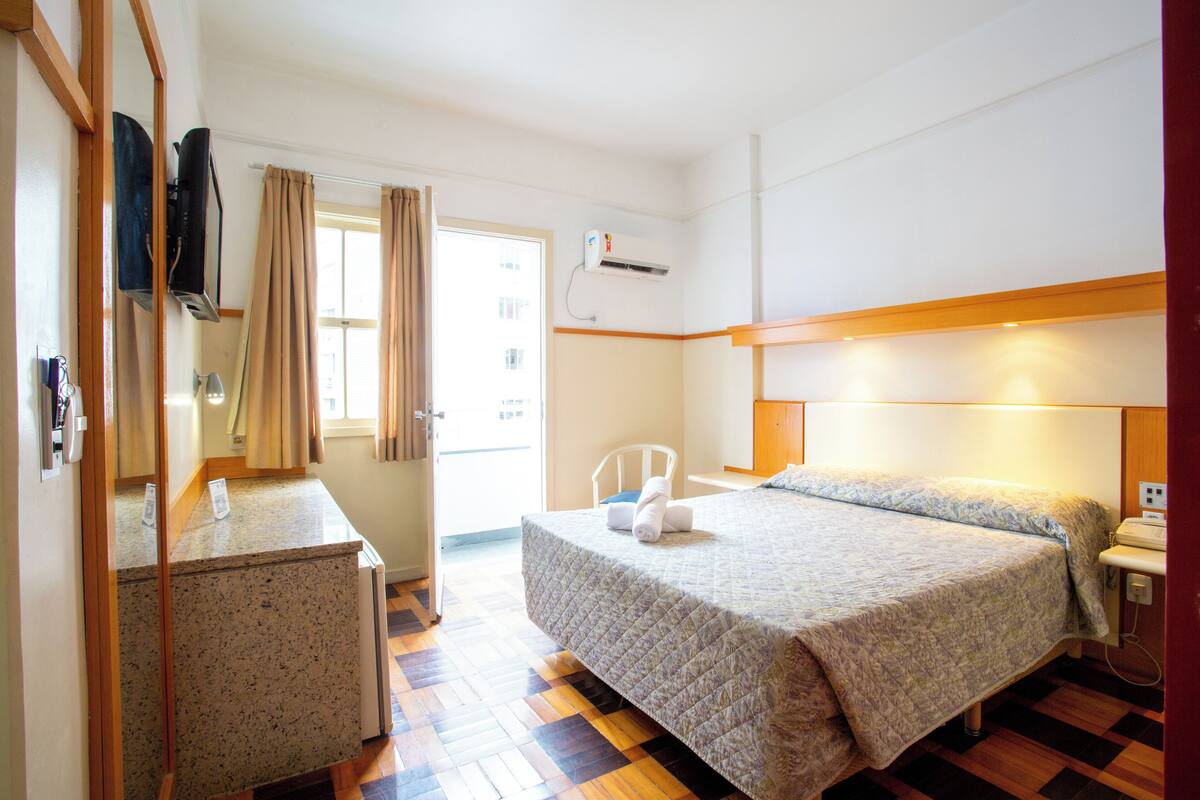 A hotel room with a bed, a desk, a chair, an air conditioning unit, and a window with curtains.