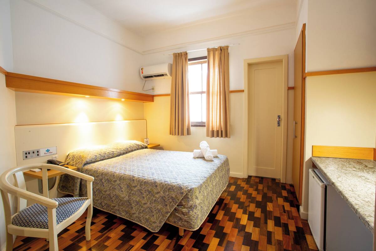 A small, neatly arranged room with a bed, a chair, a desk, and a kitchenette.