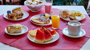 Una mesa servida con alimentos para el desayuno, incluyendo pasteles, tostadas, un vaso de jugo de naranja y una taza de café, rodeada de abundante follaje.