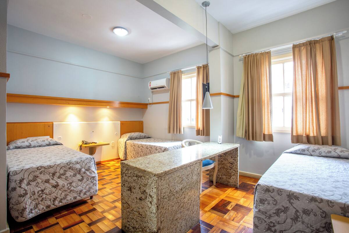 A hotel room with two beds, a desk, and a chair. There is a window with curtains, an air conditioning unit, and a lamp.