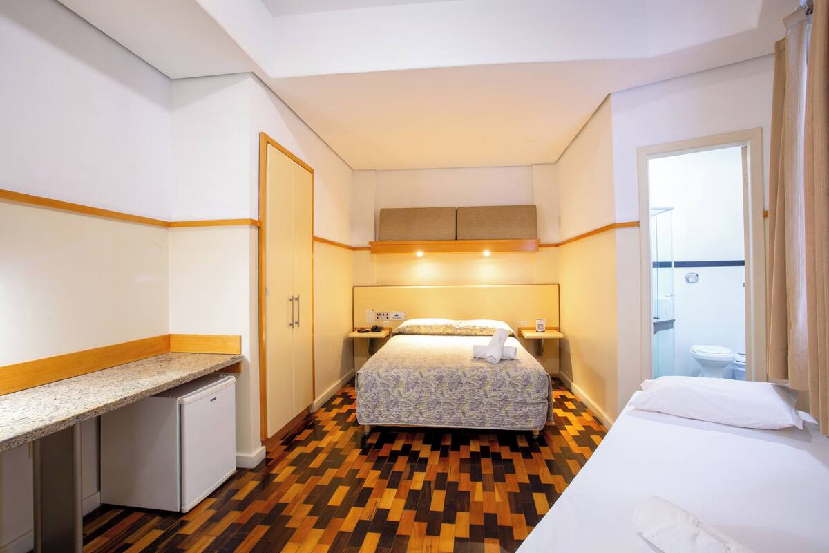 A hotel room with a bed, a small desk, a refrigerator, a bathroom, and a shower.