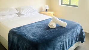 Een net opgemaakt bed met een blauwe deken, een wit kussen en een opgerolde handdoek aan de zijkant. Daarboven een plafondventilator en een raam met uitzicht op groen.