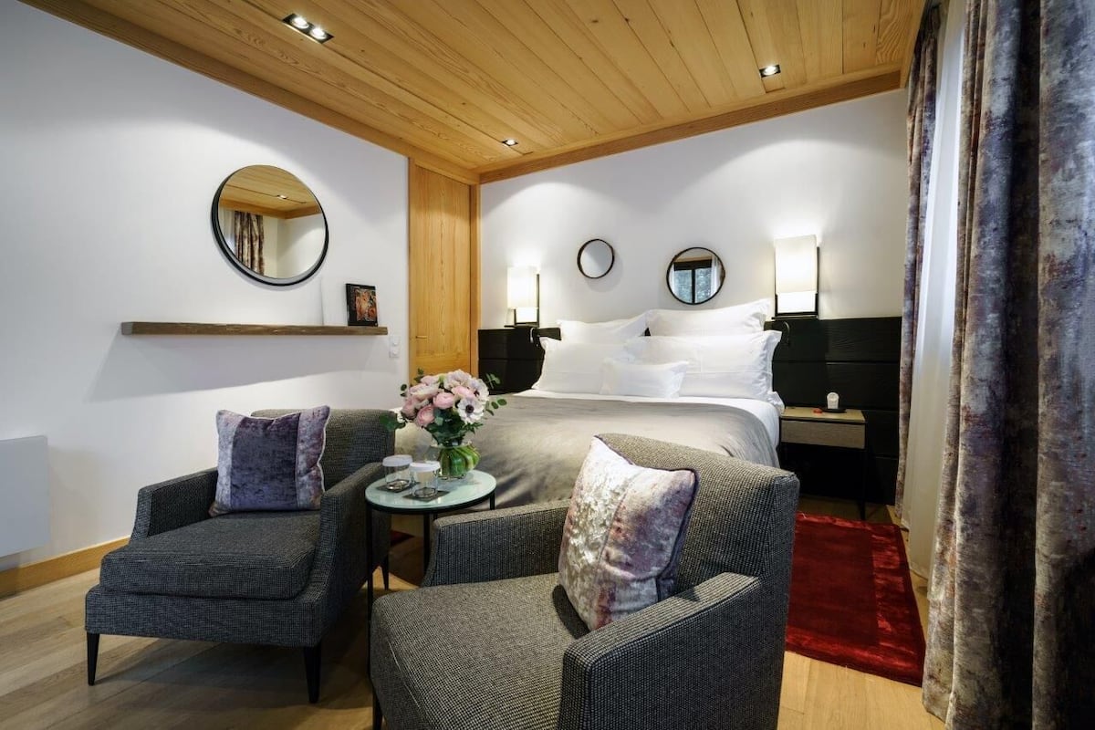 Habitación de hotel con una cama, dos sillones, una mesita con un jarrón de flores y una alfombra roja.