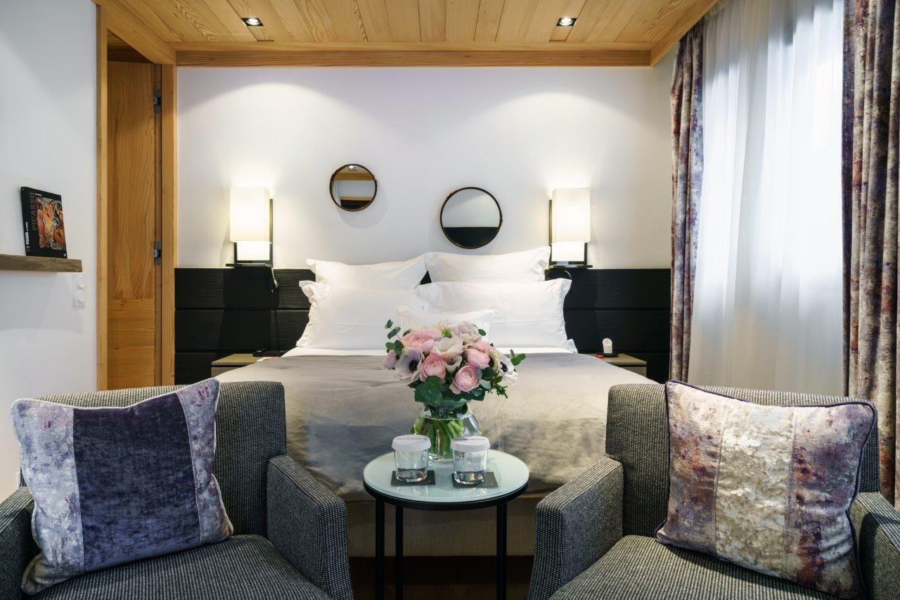 Habitación de hotel con una cama, dos mesitas de noche, una pequeña mesa redonda con velas, dos sillones y un espejo en la pared.
