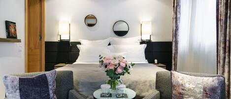Habitación de hotel con una cama, dos mesitas de noche, una pequeña mesa redonda con velas, dos sillones y un espejo en la pared.