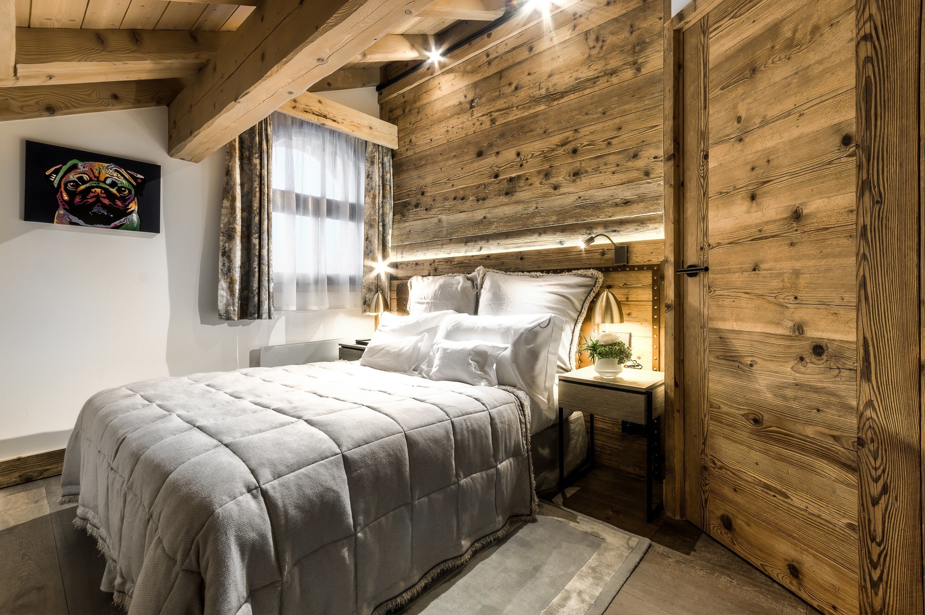 Un dormitorio con techo de madera, cama con colcha gris, mesita de noche con lámpara y puerta de madera.