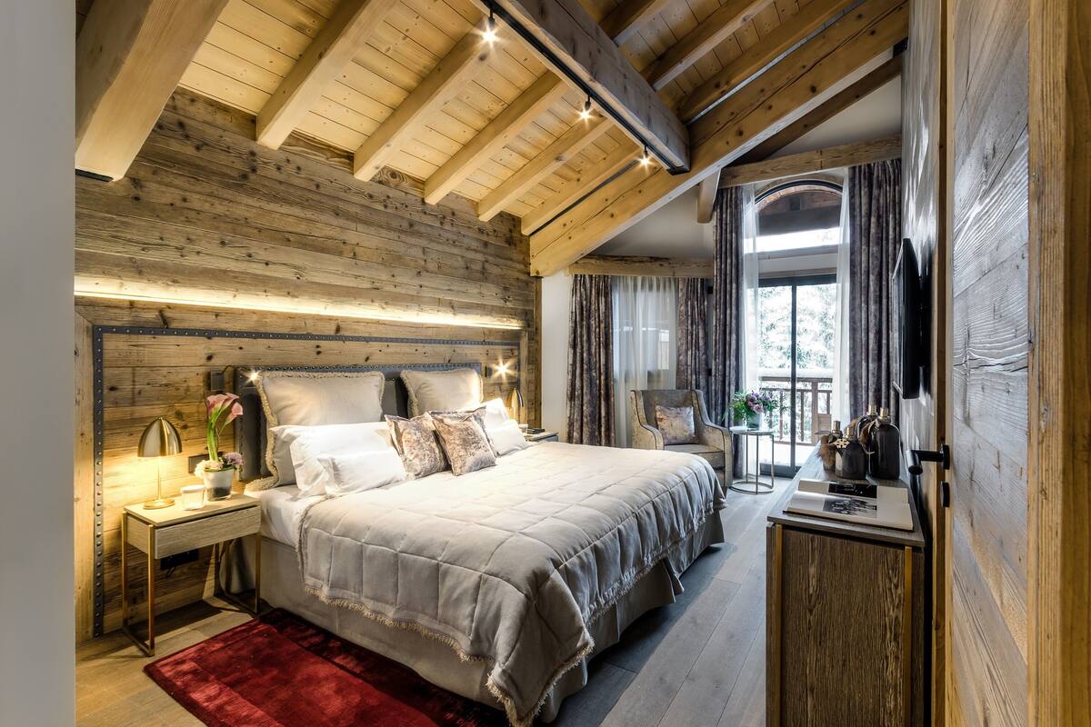 Un dormitorio con una cama grande, vigas de techo de madera, una mesita de noche con lámpara y vistas al exterior a través de una ventana.