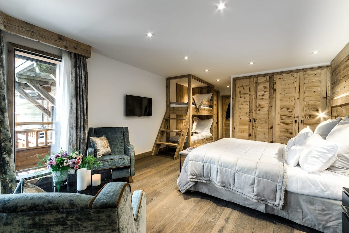 Un dormitorio con una cama litera, una escalera de madera, un sofá, un televisor montado en la pared y una ventana con cortinas.