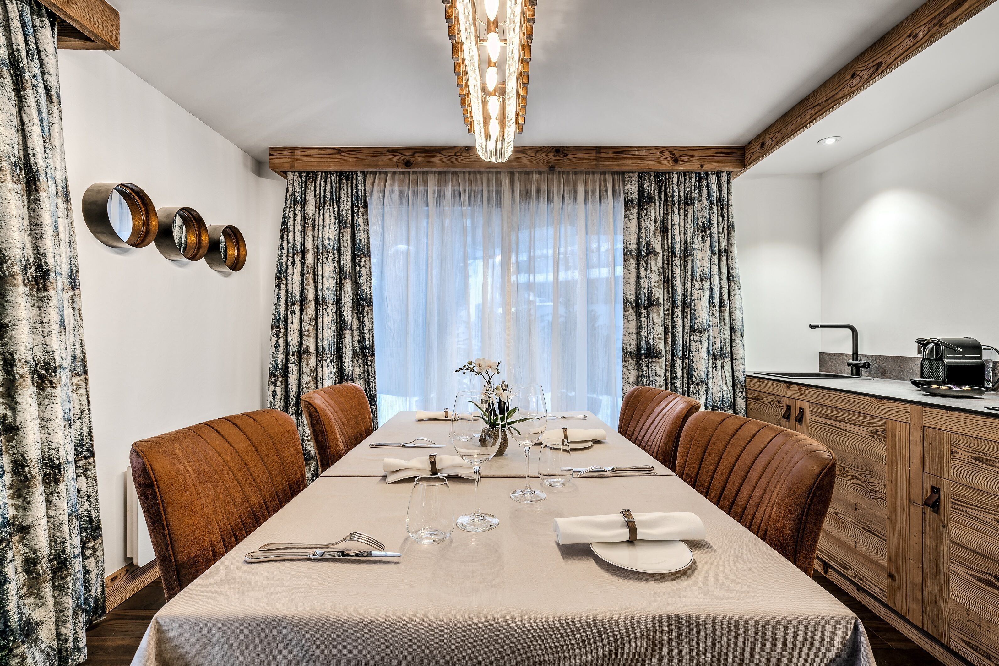 Comedor con mesa dispuesta para cuatro, sillas de madera, lámpara de araña y vistas al exterior a través de cortinas translúcidas.