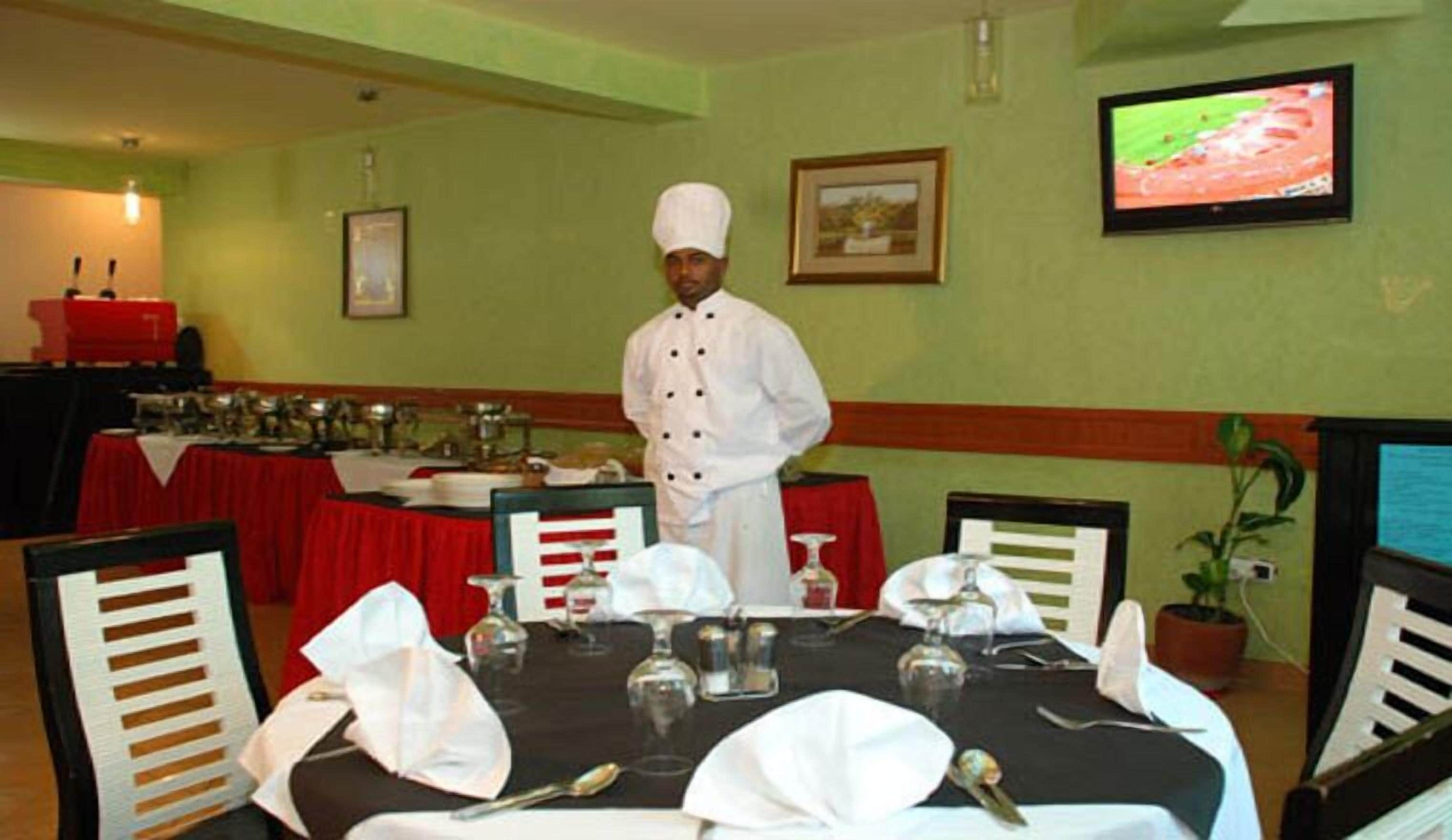 A chef standing in a dining area with a table set for a meal, a television on the wall, and a framed picture above the table.