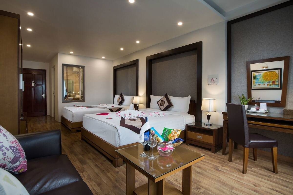 A hotel room with two beds, a desk, a chair, and a small table with a glass of water and snacks.