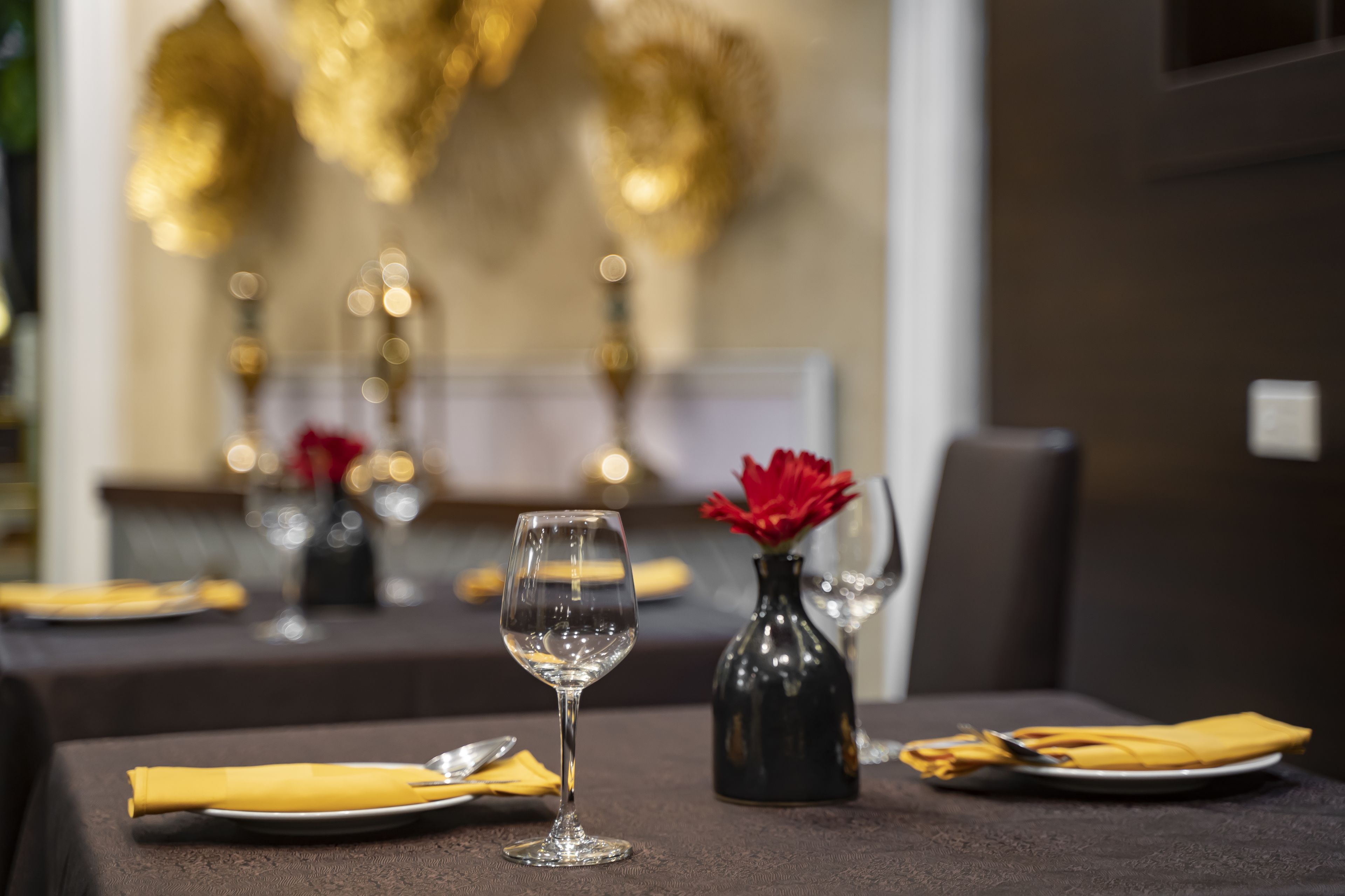 Una mesa de comedor con una copa de vino, un florero con flores rojas y servilletas amarillas dobladas con cuidado.