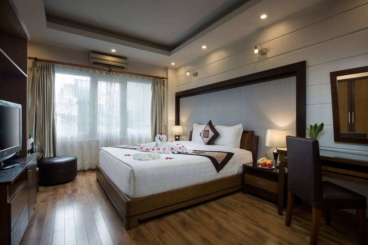 A hotel room with a bed, bedside tables, a desk, a chair, a television, and a large window with curtains.