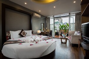 A hotel room with a large bed, a TV, a desk, and a view of the city through large windows.