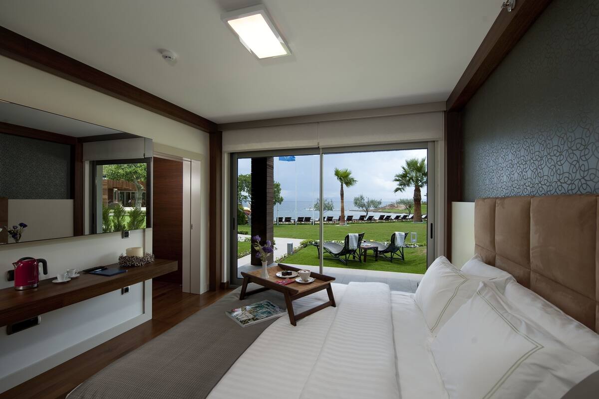 Una habitación de hotel con una cama, una mesita de noche de madera, una pequeña mesa con una bandeja, una ventana con vistas al mar y palmeras, y un espejo grande.