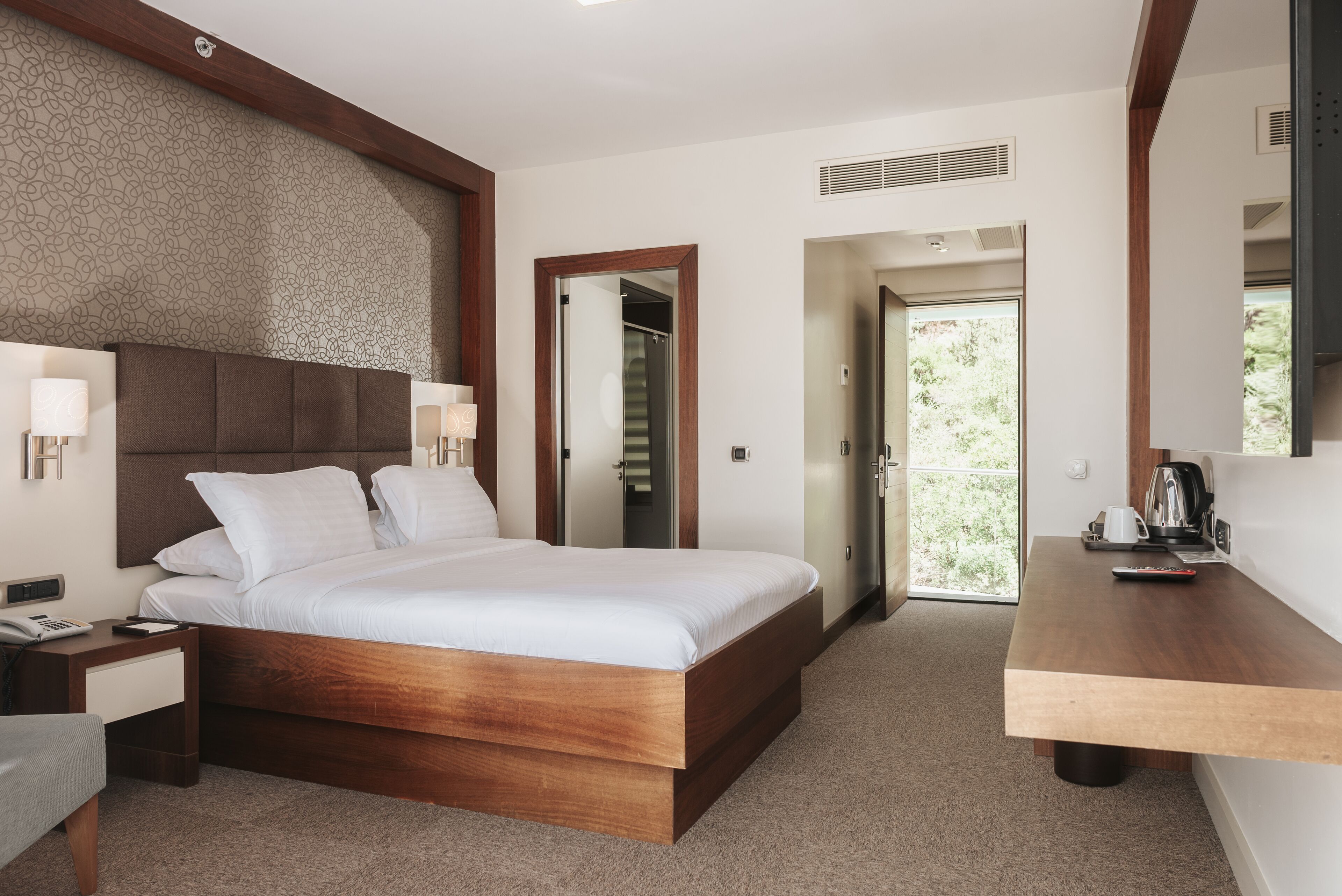 A hotel room with a wooden bed, bedside lamps, a desk with a kettle, and a view of greenery through the door.