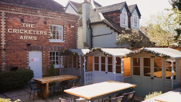 A brick building with a sign reading "The Cricketers Arms." It features outdoor seating with tables and chairs, a wooden deck, and a small wooden structure with windows. - The Cricketers Arms (Saffron Walden)