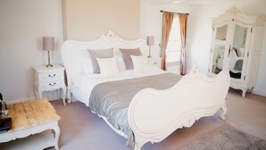 A bedroom with a white ornate bed, a nightstand with a lamp, a wooden bench, and a white wardrobe. - The Cricketers Arms (Saffron Walden)