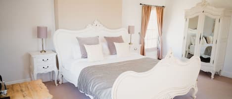 A bedroom with a white ornate bed, a nightstand with a lamp, a wooden bench, and a white wardrobe.