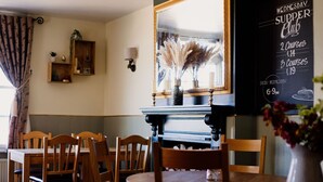 A dining area with wooden tables and chairs, a mirror, a wall-mounted shelf with decorative items, and a chalkboard menu for a supper club. - The Cricketers Arms (Saffron Walden)