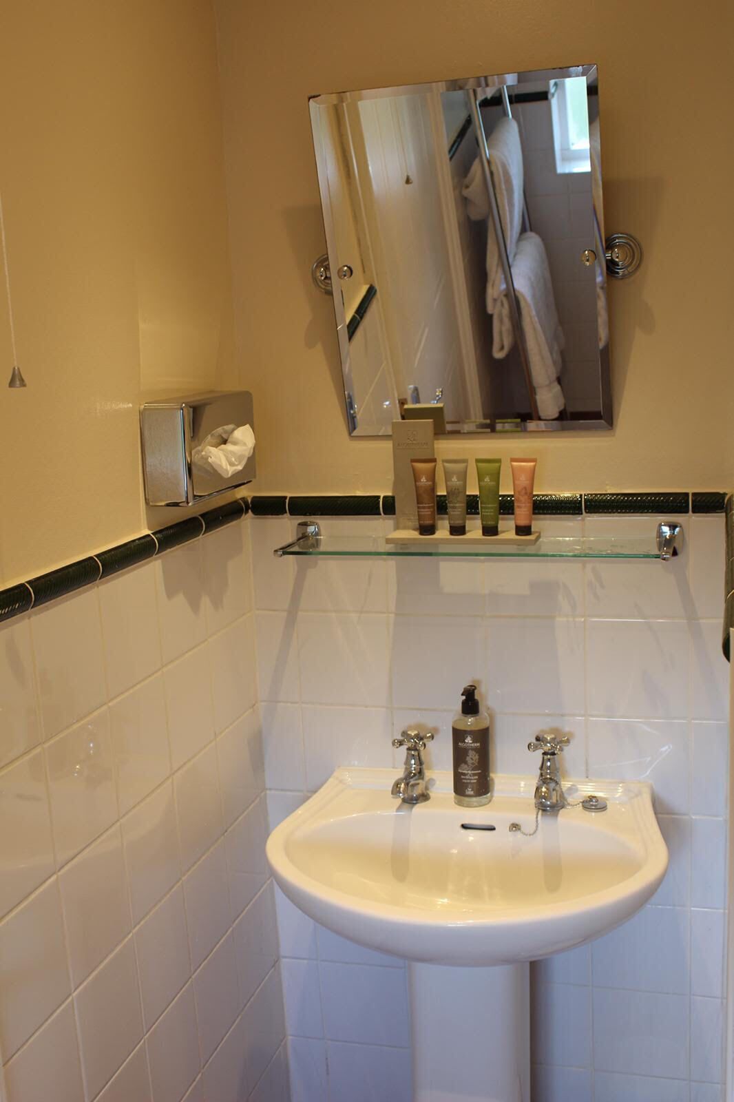 Un baño con lavamanos blanco, dos grifos, un espejo sobre el lavamanos y un estante con artículos de aseo.