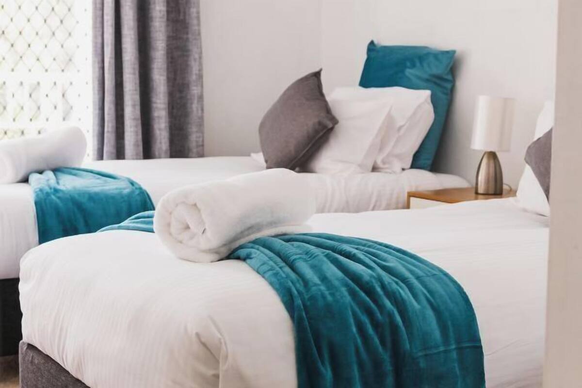 Two twin beds with white bedding and teal throw blankets, a nightstand with a lamp, and a window with curtains.