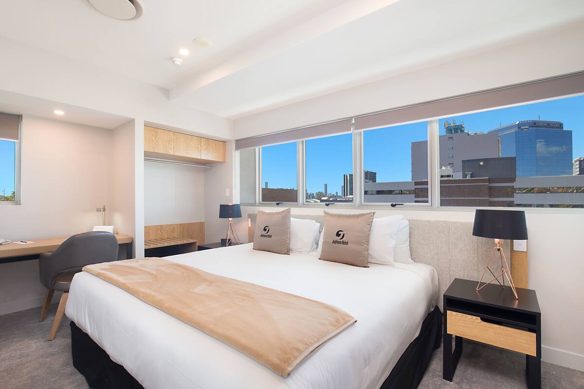 A modern hotel room with a large bed, a desk, and a view of the cityscape.