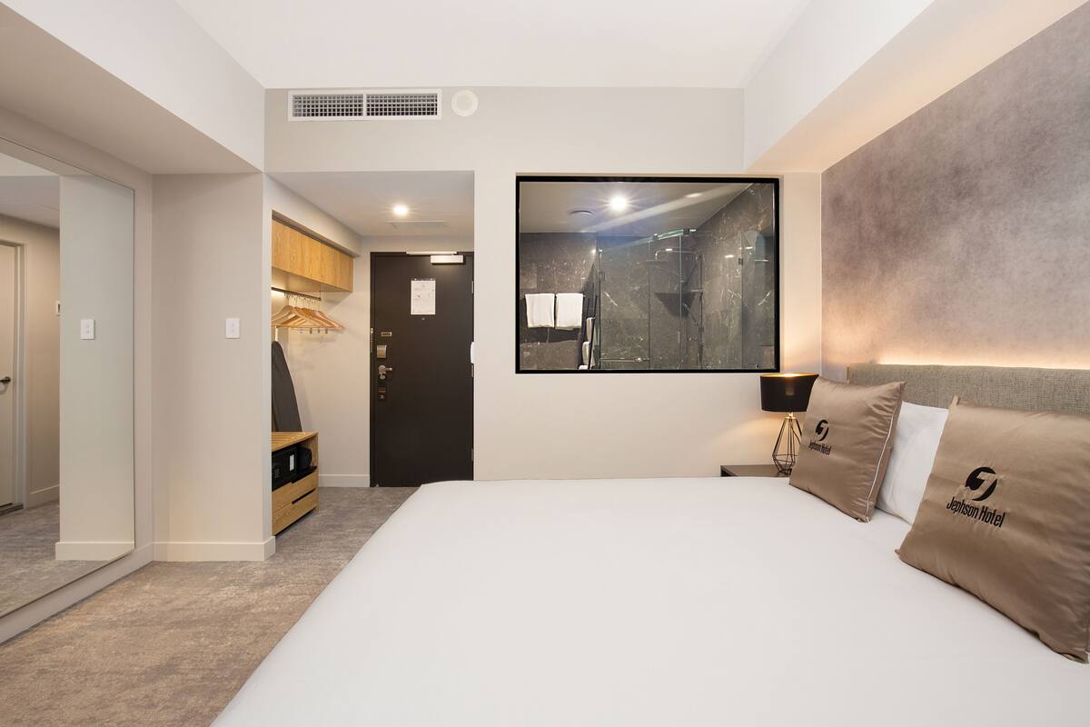 A modern hotel room with a large bed, a mounted TV, a shower area, and a wardrobe.