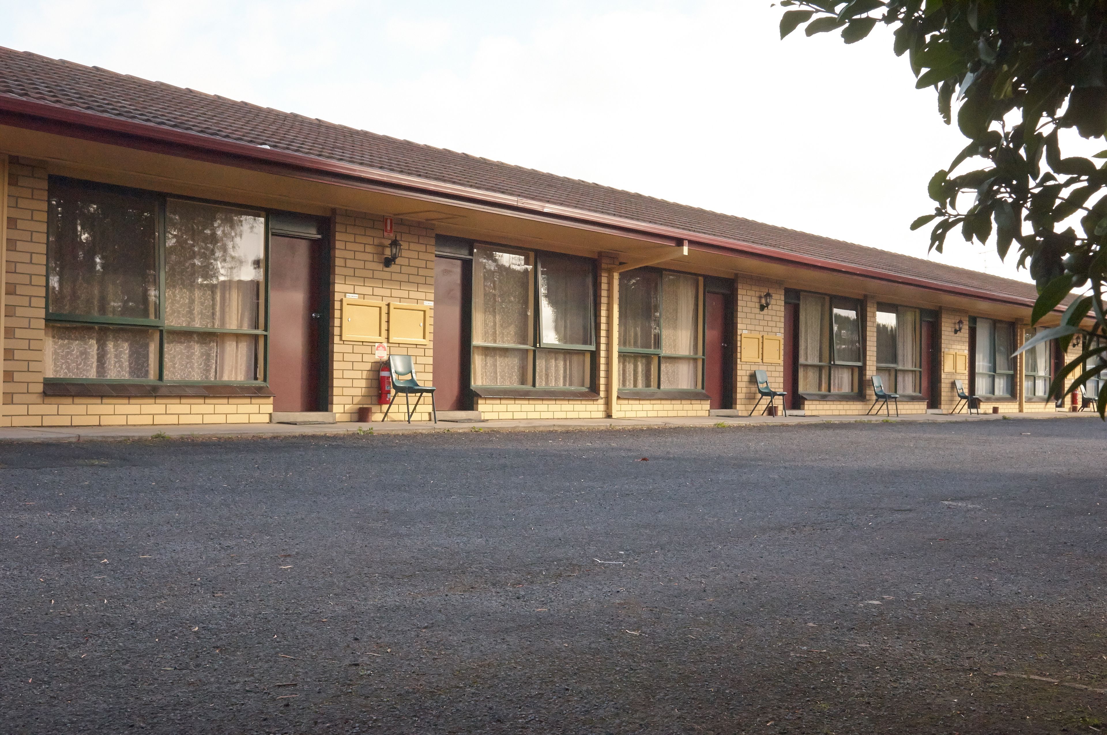 A motel with a row of rooms, each with a window and a door. There are chairs outside the rooms.