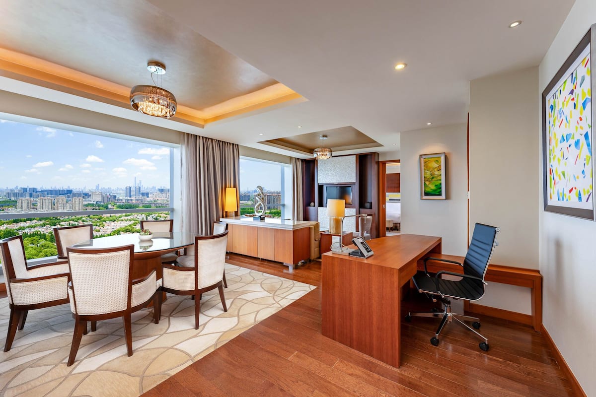 A modern hotel room with a dining area, a desk, and a view of the cityscape.